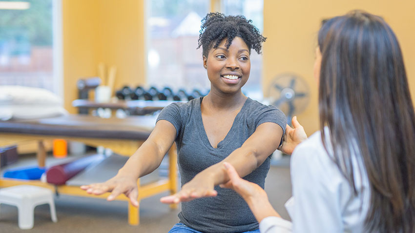 Black woman lifting her arms with the help of a physical therapist in a gym-like setting