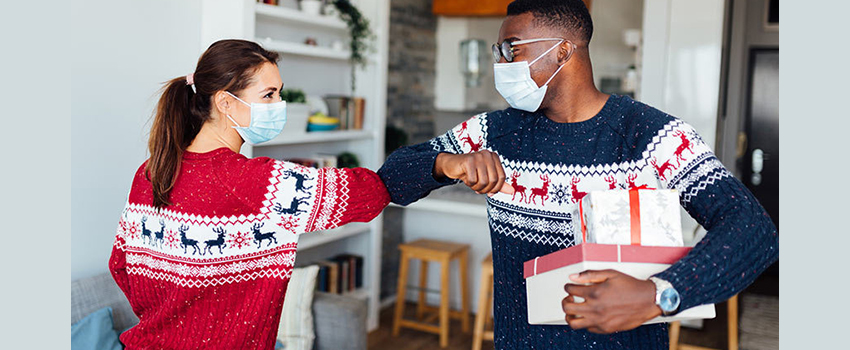 A woman and man wearing holiday sweaters and masks touch elbows