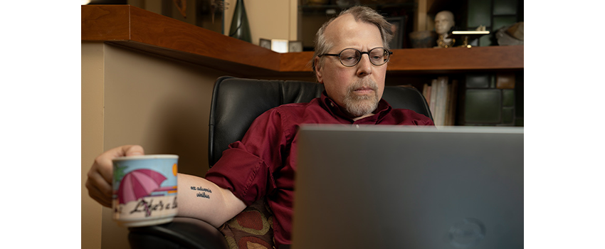 Scott Ward wears glasses and a red shirt while looking at his laptop  He holds a mug that says Life's a Beach