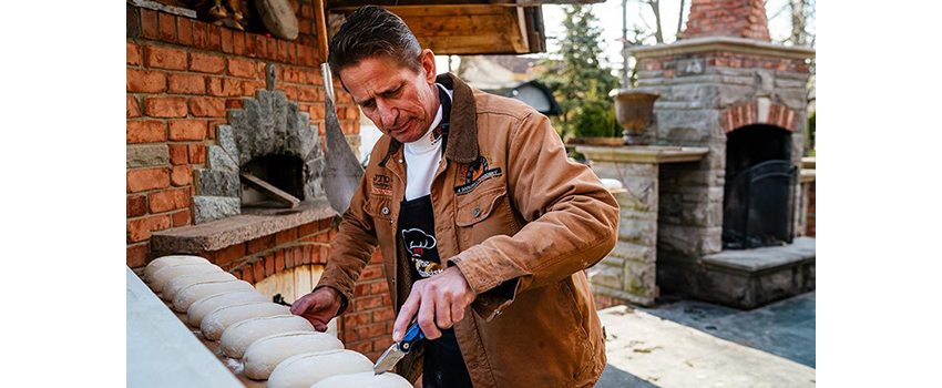 Man standing in front of outdoor oven slicing into a loaf of bread