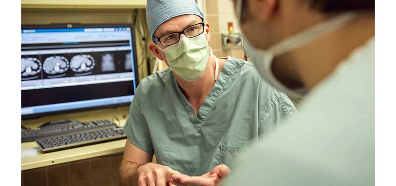 White, male doctor masked sits in front of computer screen