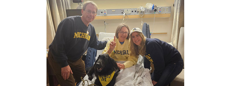 Connie Achtenberg in her hospital room with her husband, daughter and dog; all wearing maize and blue