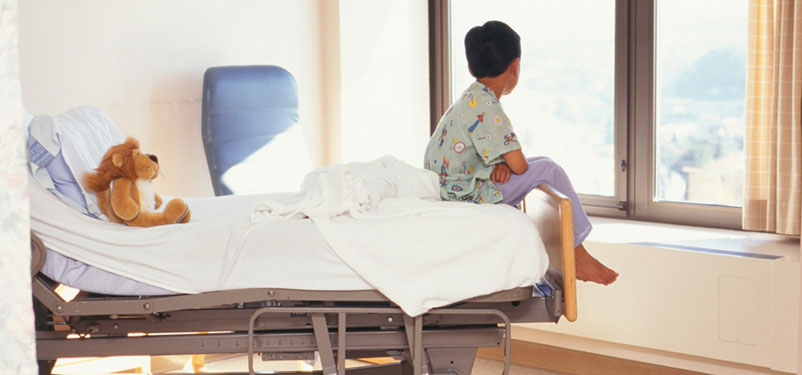 Young boy sits on the end of a hospital bed and looks out a window