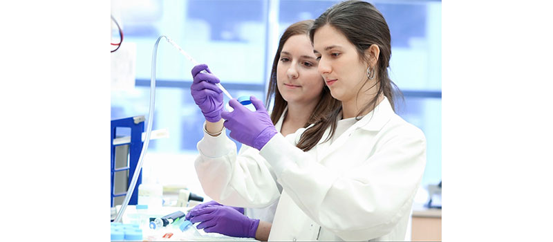 Two young white female scientists work together one holds a pipette