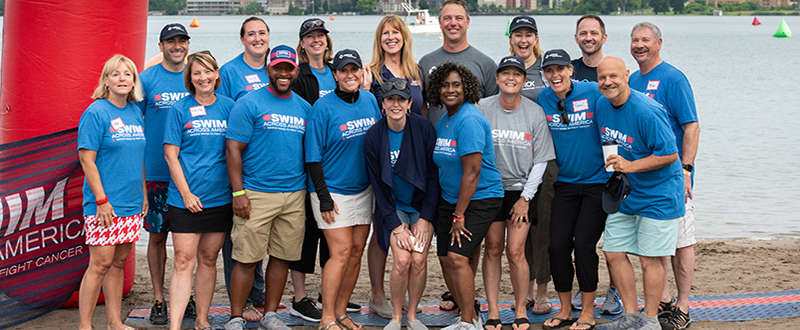 A group of men and women -- Black and white -- stand in front of the Detroit River wearing Swim Across America shirts